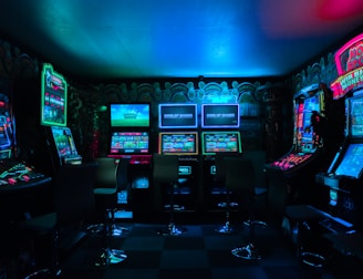 Dimly lit gaming arcade featuring a variety of brightly illuminated slot machines and video gaming screens. Neon signs and colorful displays create a vibrant atmosphere. The arrangement of gaming machines suggests an inviting space for entertainment, with chairs positioned for players.