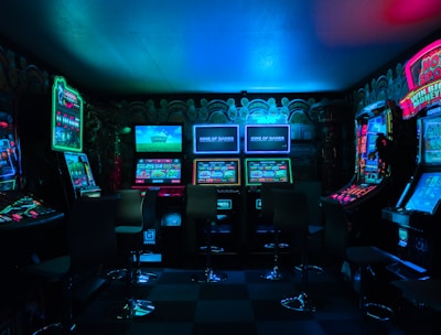 Dimly lit gaming arcade featuring a variety of brightly illuminated slot machines and video gaming screens. Neon signs and colorful displays create a vibrant atmosphere. The arrangement of gaming machines suggests an inviting space for entertainment, with chairs positioned for players.