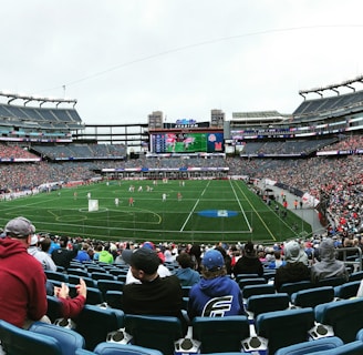 A large sports stadium filled with spectators. The field is green with white markings, and players in red and white uniforms are actively engaged in a game. The stands are packed with people wearing various colored clothing, and some sections of the crowd are more densely populated. Digital screens and advertisements are visible around the stadium, adding to the vibrant atmosphere.