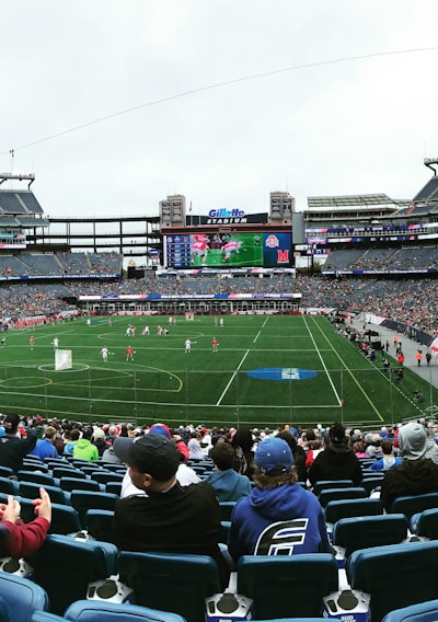 A large sports stadium filled with spectators. The field is green with white markings, and players in red and white uniforms are actively engaged in a game. The stands are packed with people wearing various colored clothing, and some sections of the crowd are more densely populated. Digital screens and advertisements are visible around the stadium, adding to the vibrant atmosphere.