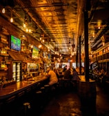 A dimly lit bar interior with a warm ambiance, featuring several television screens displaying sports broadcasts above a wooden counter. Patrons are seated at the bar and tables, engaged in conversation. The ceiling is ornate with decorative panels and hanging light fixtures add a cozy glow to the scene.