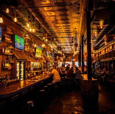 A dimly lit bar interior with a warm ambiance, featuring several television screens displaying sports broadcasts above a wooden counter. Patrons are seated at the bar and tables, engaged in conversation. The ceiling is ornate with decorative panels and hanging light fixtures add a cozy glow to the scene.