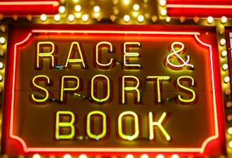 Bright neon lights on a marquee sign display the words 'RACE & SPORTS BOOK'. The sign is surrounded by rows of yellow and white lights against a red background, evoking a vibrant, energetic atmosphere typically associated with casinos or entertainment venues.