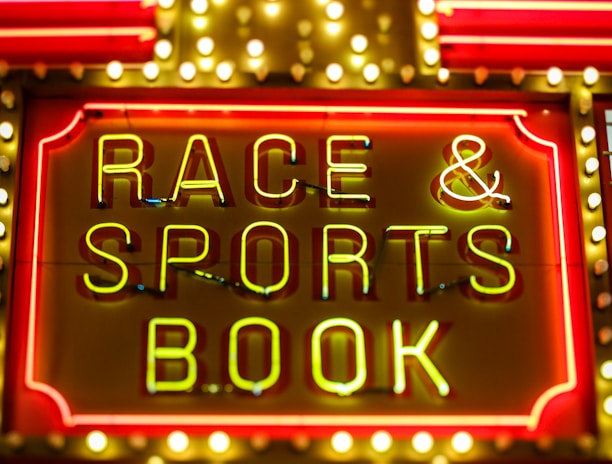 Bright neon lights on a marquee sign display the words 'RACE & SPORTS BOOK'. The sign is surrounded by rows of yellow and white lights against a red background, evoking a vibrant, energetic atmosphere typically associated with casinos or entertainment venues.