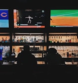 A bar counter with several people sitting in front of it is shown. Above the counter, there are multiple television screens displaying sports broadcasts, including football and baseball. Behind the counter, various bottles of alcohol are neatly arranged.