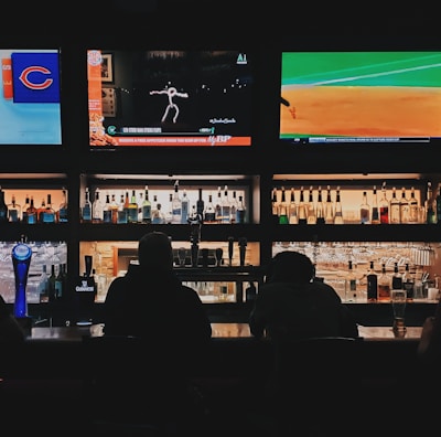 A bar counter with several people sitting in front of it is shown. Above the counter, there are multiple television screens displaying sports broadcasts, including football and baseball. Behind the counter, various bottles of alcohol are neatly arranged.