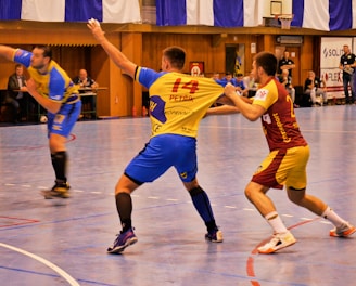 A dynamic indoor sports scene with players in motion on a court. Two athletes in blue and yellow uniforms are engaged in an intense handball game, with one player gripping the other's jersey. The background shows other players, several seated people, and advertisements on the walls.