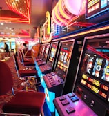 A brightly lit casino interior featuring a row of colorful slot machines. The neon lights create a vibrant atmosphere, with patterned carpeting adding to the decor. Stools are aligned neatly in front of each machine.