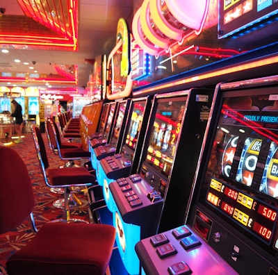 A brightly lit casino interior featuring a row of colorful slot machines. The neon lights create a vibrant atmosphere, with patterned carpeting adding to the decor. Stools are aligned neatly in front of each machine.