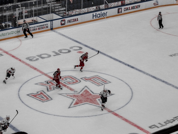 An ice hockey game is underway in an indoor arena with players actively engaged in the sport. Spectators are seated in the stands, while some areas appear sparse. The rink features a large logo at center ice, and electronic advertisements surround the perimeter.