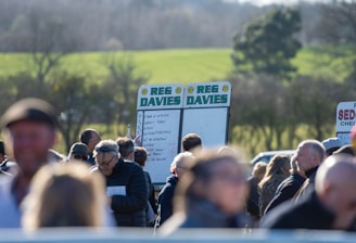 A group of people gathered outdoors with two large betting boards in the background labeled 'REG DAVIES'. The setting appears to be a countryside location with trees and fields visible in the distance. The crowd consists of individuals dressed in jackets and hats, engaging in conversation.