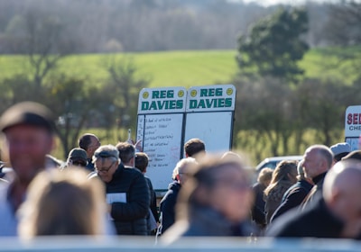 A group of people gathered outdoors with two large betting boards in the background labeled 'REG DAVIES'. The setting appears to be a countryside location with trees and fields visible in the distance. The crowd consists of individuals dressed in jackets and hats, engaging in conversation.