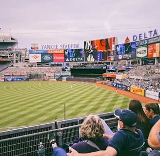 A baseball stadium filled with spectators watching a game on a neatly maintained grass field. The stadium features large advertising screens displaying various logos and brands. Fans sitting in the foreground are dressed casually, enjoying the sporting event.