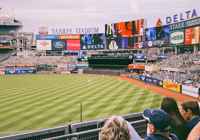 A baseball stadium filled with spectators watching a game on a neatly maintained grass field. The stadium features large advertising screens displaying various logos and brands. Fans sitting in the foreground are dressed casually, enjoying the sporting event.
