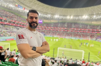 A person stands in a stadium filled with spectators during a sporting event. The grass field and goalposts are visible in the background. The individual is wearing a white jersey with a sports logo and is smiling at the camera.