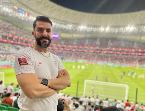 A person stands in a stadium filled with spectators during a sporting event. The grass field and goalposts are visible in the background. The individual is wearing a white jersey with a sports logo and is smiling at the camera.