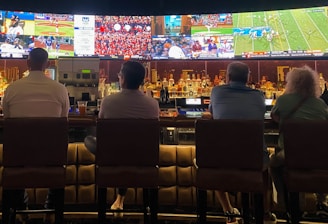 A group of people sit at a bar with their backs to the camera, watching multiple sports games displayed on large screens. The bar counter is lined with various bottles of liquor, and the atmosphere suggests a casual and relaxed social setting.