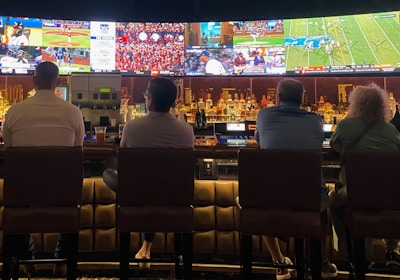 A group of people sit at a bar with their backs to the camera, watching multiple sports games displayed on large screens. The bar counter is lined with various bottles of liquor, and the atmosphere suggests a casual and relaxed social setting.