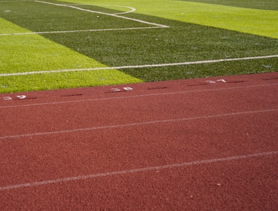 A sports field with vivid green grass divided into sections by white lines in the background. In the foreground, a track with a reddish-brown surface is marked with numbers and lines.
