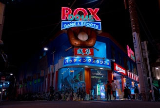 A vibrant, neon-lit facade of a gaming and sports establishment at night, featuring a large glove holding a baseball above the entrance. The building is adorned with colorful signs in both English and Japanese, with various advertisements and an illuminated sign that says 'ROX DOME'. Several people are walking towards the entrance, and bicycles are parked along the side.