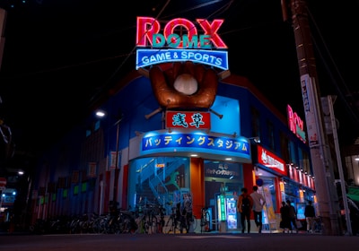 A vibrant, neon-lit facade of a gaming and sports establishment at night, featuring a large glove holding a baseball above the entrance. The building is adorned with colorful signs in both English and Japanese, with various advertisements and an illuminated sign that says 'ROX DOME'. Several people are walking towards the entrance, and bicycles are parked along the side.