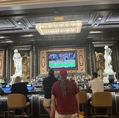 Several people are seated at a luxurious bar, with a large TV screen showing a sports game. The bar is elegantly decorated with ornate details and sculptures. The ceiling has a large, rectangular, crystal chandelier.
