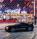 A sleek black sports car is parked in front of an illuminated arena entrance, with a large sign bearing the name of a famous beverage brand. The building features a modern, geometric facade with bright red and blue lights, creating a vibrant and lively atmosphere.