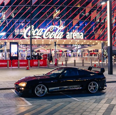A sleek black sports car is parked in front of an illuminated arena entrance, with a large sign bearing the name of a famous beverage brand. The building features a modern, geometric facade with bright red and blue lights, creating a vibrant and lively atmosphere.